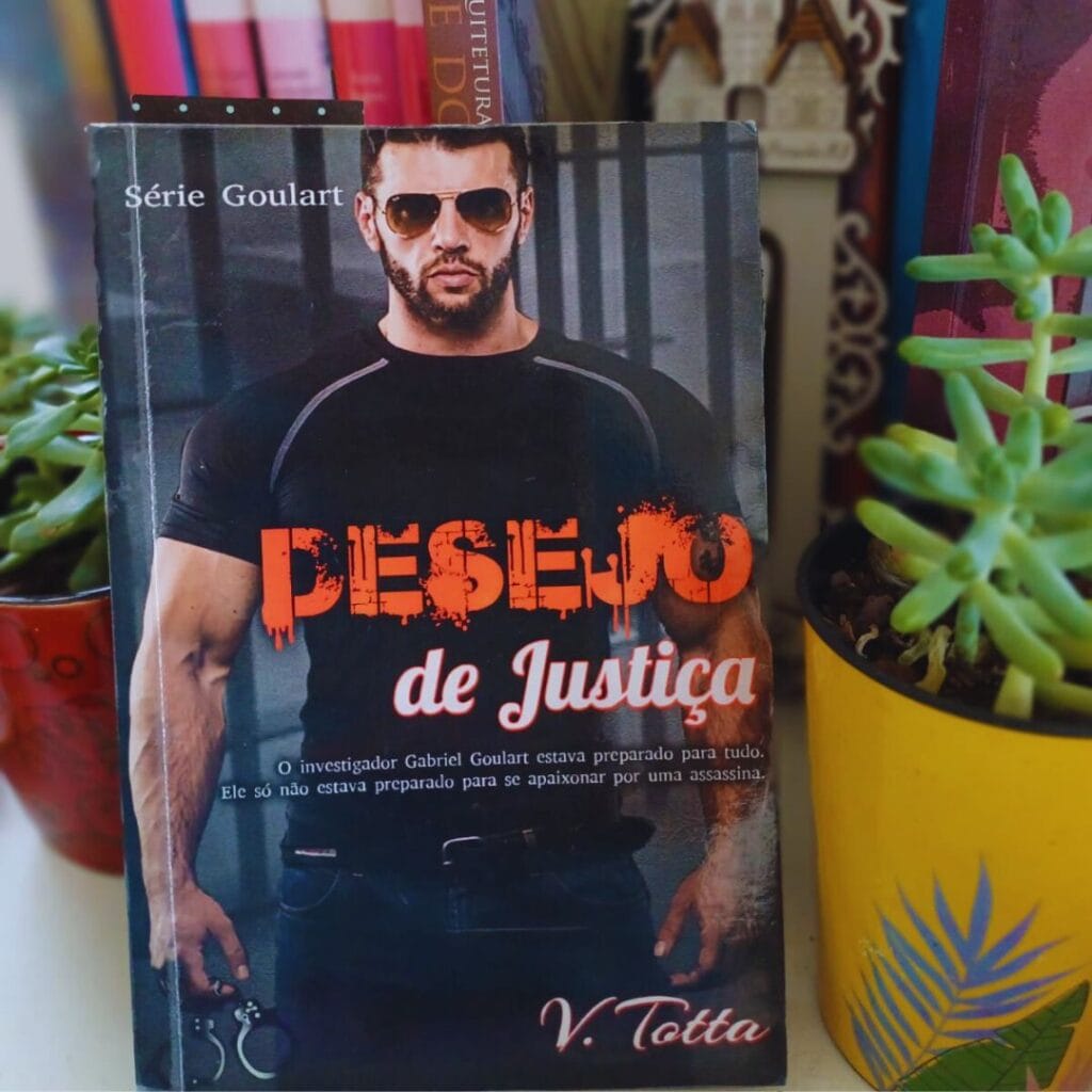 Capa do livro Desejo de Justiça, da autora V. Totta.
O livro está numa superfície branca e apoiado em outros livros de diversos formatos e tamanhos, ao lado dele estão duas canecas de cerâmica, nas cores vermelha e amarela, contendo suculentas. Na capa do livro predominam as cores preto, branco e vermelho. Em destaque há a imagem de um homem branco, alto, forte e de semblante sério, com barba escura cerrada, cabelo escuro e curto e óculos escuros. Ele usa uma camiseta preta e calça jeans escura, na mão direita ele segura um par de algemas. No centro da capa está a palavra "Desejo" em letras grandes e vermelhas, que escorrem como de fosse sangue e "de justiça" em letras brancas em tamanho menor.
